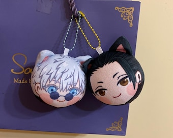 May include: Two plush character keychains, one with white hair and sunglasses, the other with black hair and cat ears. Both have smiling faces and are attached to a gold chain.