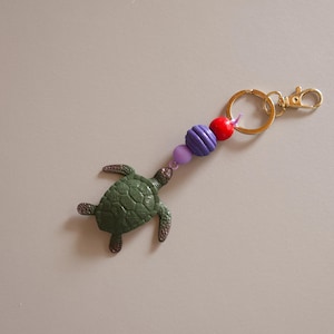 Könnte beinhalten: Ein grüner Schildkröten-Schlüsselanhänger mit einem goldfarbenen Verschluss und Ring. Der Schlüsselanhänger hat eine Kette aus lila, roten und blauen Perlen. Die Schildkröte hat einen strukturierten Panzer und ist aus dunkelgrünem Material.
