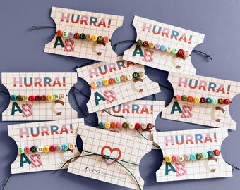 May include: A collection of bracelets on rectangular display cards. Each card features a grid pattern, the text HURRA!, and the letters A, B, and C. The bracelets consist of colourful round beads spelling the word SCHULKIND on a dark string.