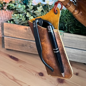 May include: A handcrafted leather sunglasses case in shades of brown and tan, holding black sunglasses and a black pen. The case has a loop for hanging and is held up by a wooden hand.