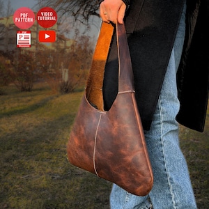 Puede incluir: Un bolso de hombro de cuero marrón con una correa larga. El bolso está usado y tiene un aspecto desgastado. El bolso lo lleva una persona que lleva vaqueros y una chaqueta negra.