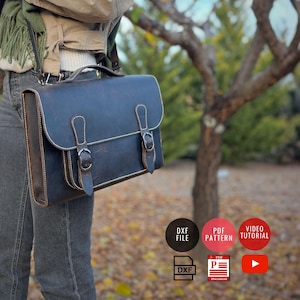 May include: A dark blue leather satchel with a rectangular shape, featuring a top handle and two buckle closures. The bag has brown trim and white stitching. The bag is being carried over the shoulder. The background includes a tree and icons for DXF file, PDF pattern, and video tutorial.