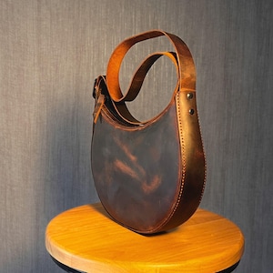 May include: A brown leather satchel bag with a curved shape and a long strap. The bag has a distressed look and is sitting on a wooden surface.