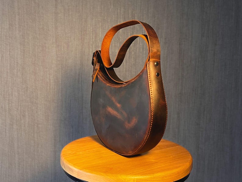 May include: A brown leather satchel bag with a curved shape and a long strap. The bag has a distressed look and is sitting on a wooden surface.