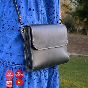 Può includere: Borsa a tracolla in pelle nera con chiusura a patta e cuciture bianche. La borsa ha una tracolla lunga e regolabile. Lo sfondo mostra un vestito blu e erba verde.