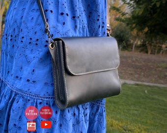 May include: A black leather crossbody bag with a flap closure and white stitching. The bag has a long, adjustable strap. The background shows a blue dress and green grass.