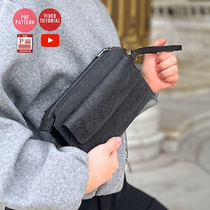 Puede incluir: Clutch negra con un estampado de cachemira. El clutch tiene un compartimento principal con cremallera y un bolsillo frontal con solapa. Se adjunta una pequeña correa de muñeca. El clutch lo sostiene una persona que lleva una chaqueta gris. Iconos de patrón PDF y tutorial de vídeo en el fondo.