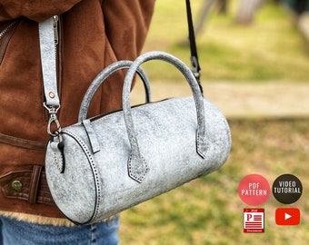 May include: A gray leather cylindrical handbag with a top handle and a shoulder strap. The bag has a zipper closure and decorative stitching. The bag is being held by a person wearing a brown jacket and blue jeans. The image also includes text that says "PDF PATTERN" and "VIDEO TUTORIAL".