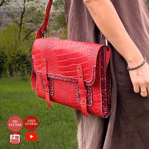 Puede incluir: Bolso de cuero rojo con textura de piel de cocodrilo, con cierre de solapa y correa ajustable para el hombro. El bolso tiene dos correas con hebilla en la parte delantera y se lleva al hombro. El fondo muestra un campo verde y árboles.