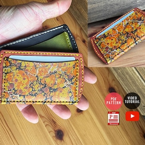 May include: A leather card holder with a marbled pattern in shades of orange, yellow, and black. The card holder is folded in half and has a stitched edge. The card holder is being held in a hand.