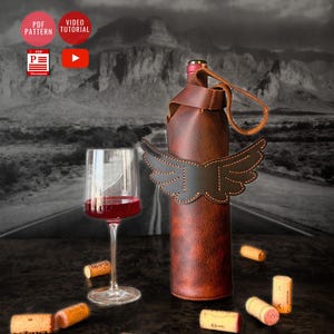 May include: A brown leather wine bottle carrier with a wing design. The carrier is on a table with wine corks scattered around it. A glass of red wine is in the foreground.