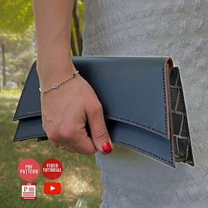 Könnte beinhalten: Dunkelblaue Leder-Clutch mit geometrischem Muster. Die Clutch wird von einer Person gehalten, die ein goldenes Armband mit blauen und schwarzen Akzenten trägt. Der Hintergrund ist eine verschwommene Szene im Freien.