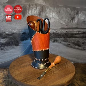 Puede incluir: Un portaherramientas de cuero hecho a mano en negro y naranja. El soporte contiene tijeras y otras herramientas. Una herramienta de marroquinería descansa sobre una superficie de madera. El soporte mide unos 15 cm de alto.