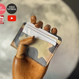 May include: A camouflage leather card holder with a stitched edge, holding several white cards. The card holder is being held by a wooden hand.