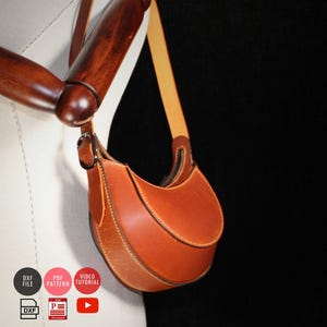 May include: A brown leather saddlebag with a curved design and a long strap. The bag has a zipper closure and is attached to a wooden mannequin arm. The bag is a rich, warm brown color with visible stitching.