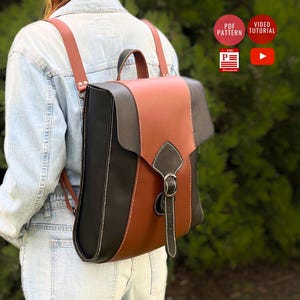 May include: A handmade leather backpack in black, brown, and tan with a buckle closure. The bag has adjustable brown straps and a top handle. The design includes white stitching and a flap closure. The backpack is shown from the back.