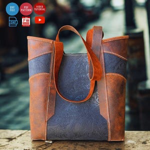 May include: A brown and blue leather tote bag with a paisley pattern. The bag has a rectangular shape with a handle and is made of different shades of brown and blue leather. The bag is sitting on a wooden surface.