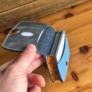 May include: A grey and blue leather card holder with a snap closure. The holder is open and shows multiple cards inside.