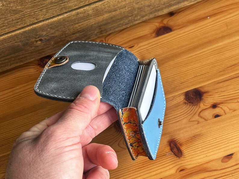May include: A grey and blue leather card holder with a snap closure. The holder is open and shows multiple cards inside.