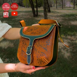 Puede incluir: Un bolso de cuero marrón con un patrón floral detallado y ribetes verdes. El bolso tiene un cierre de hebilla y una correa para el hombro. La imagen también incluye iconos para un patrón PDF y un tutorial en vídeo.