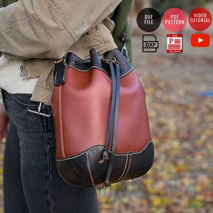 May include: A brown and black leather bucket bag with a drawstring closure. The bag has a rounded shape with a black base and top trim. The bag is worn over the shoulder with a long strap. The bag is approximately 10 inches tall.