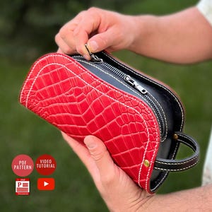 Könnte beinhalten: Eine rote, strukturierte Kulturtasche aus Leder mit schwarzem Innenraum und weißen Nähten. Die Tasche hat einen Reißverschluss und einen kleinen Griff. Die Tasche wird von einer Person gehalten.