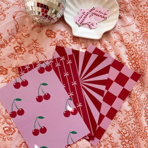 May include: Set of five pink and red patterned paper sheets. One sheet features a repeating pattern of cherries on a pink background. Another sheet has a red and pink checkered pattern. The remaining three sheets have different red and pink geometric patterns.