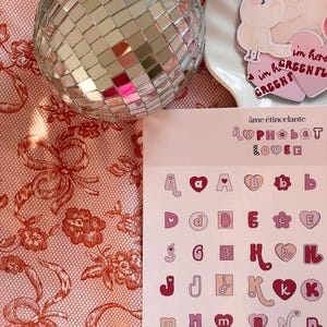 May include: Pink and white alphabet stickers with hearts and flowers. The stickers are on a pink background with the text "&acirc;me &eacute;tincelante alphabet lover" at the top.