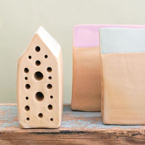 Könnte beinhalten: Drei dekorative Keramik-Häuser in einer neutralen Farbpalette. Ein hausförmiges Stück hat mehrere kreisförmige Öffnungen. Die anderen beiden sind rechteckig mit pastellfarbenen Oberteilen. Sie stehen auf einer verwitterten Holzoberfläche.