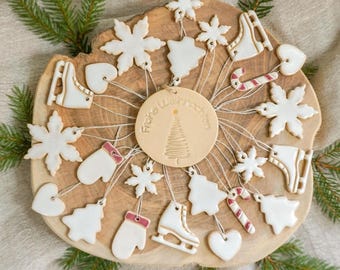 May include: A wooden Christmas ornament display featuring various white and red-accented holiday shapes, including snowflakes, mittens, and ice skates. The ornaments are arranged on a wooden slice, with a central disc that reads "Frohe Weihnachten".
