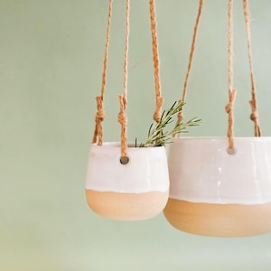 Könnte beinhalten: Zwei hängende Keramik-Pflanzgefäße, eines klein und eines groß, mit zweifarbigem Design. Die obere Hälfte ist weiß und die untere Hälfte ist hellbraun. Sie sind an Naturseilen aufgehängt und enthalten Grünzeug.