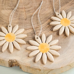 Könnte beinhalten: Drei Ornamenten in Form von Gänseblümchen mit gelben Zentren und cremefarbenen Blütenblättern, auf weißen Kordeln aufgereiht. Die Ornamente liegen auf einem Holzschneidebrett mit natürlichem Holzmaserungsmuster. Die Gänseblümchen sind eine charmante, handgefertigte Dekoration.