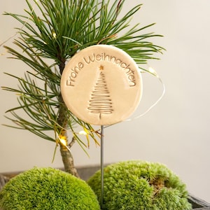 Könnte beinhalten: Ein runder, beigefarbener Keramik-Anhänger mit dem Text "Frohe Weihnachten" und einem stilisierten Weihnachtsbaum-Design. Der Anhänger ist an einer kleinen, grünen Topfpflanze befestigt.