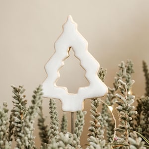 Könnte beinhalten: Ein weißer Keramik-Weihnachtsbaumschmuck mit einem Ausschnittmuster, umgeben von grünem Laub und Lichterketten.