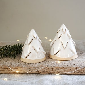 Könnte beinhalten: Zwei weiße Keramik-Weihnachtsbaumschmuckstücke mit einem strukturierten Design. Die Schmuckstücke liegen auf einer Holzoberfläche mit weißen Lichterketten.