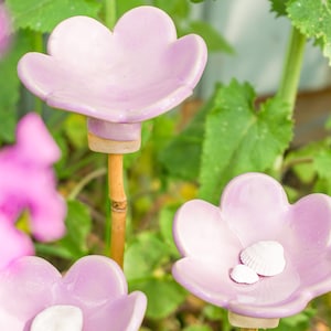 Könnte beinhalten: Drei hellviolette Gartenstecker in Form von Blumen aus Keramik mit Bambusstiel. Eine Blume enthält zwei kleine weiße Muscheln. Der Hintergrund umfasst grüne Blätter und rosa Blüten.