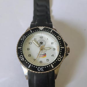 Könnte beinhalten: Eine silberfarbene Armbanduhr mit schwarzer Lünette und schwarzem Kautschukarmband. Das Zifferblatt ist weiß mit schwarzen Ziffern und Zeigern. Die Uhr zeigt das Logo des Deutschen Fußball-Bundes und eine kleine deutsche Flagge.