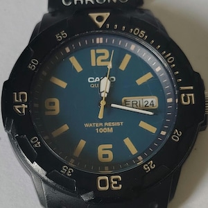 May include: Black Casio watch with a teal face, gold numerals, and a day/date display. The watch features a black bezel with minute markers and the text "WATER RESIST 100M". The watch band is black.