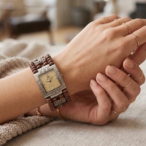 May include: A rectangular silver-tone watch with a champagne-coloured face featuring the text "Phoebe" and "Quartz." The watch band consists of multiple strands of small brown wooden beads interspersed with decorative silver-tone metal accents.