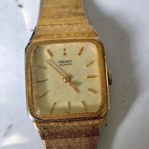 May include: A gold-tone Seiko Quartz wristwatch featuring a square face with a light-colored dial, thin gold hour markers, and matching gold hands. The watch includes a textured gold-tone metal link bracelet and a small crown on the right side.