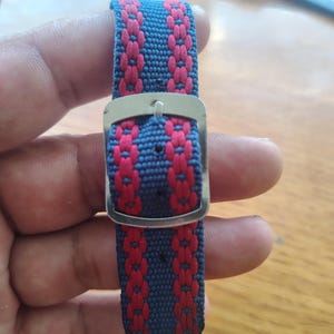 May include: A woven watch strap featuring a blue base with two vertical rows of pink floral-like patterns. The strap includes a silver-toned metal buckle with a central pin and multiple adjustment holes for securing the band around a wrist.