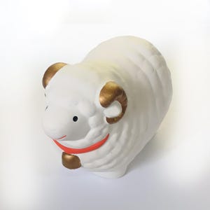 May include: A white ceramic sheep figurine with gold-coloured horns and hooves. The sheep has a red collar and a smiling mouth. The figurine is a decorative item.