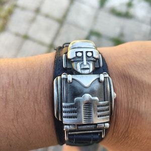May include: A black leather cuff bracelet featuring a large, silver-toned metal centrepiece shaped like a stylised robot figure. The robot design includes a rounded head, rectangular torso, and detailed mechanical lines, securely attached to the leather band.