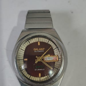 Puede incluir: Un reloj de pulsera automático en tono plateado con una esfera circular de color marrón oscuro, manecillas doradas y una ventana de día y fecha que muestra 5 FRI 26. La esfera incluye el texto GALANT AUTOMATIC y 21 JEWELS con un logo verde.