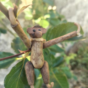 May include: A small monkey figurine crafted from natural wood pieces, featuring a rounded head with dark eyes, a prominent snout, and a light-colored neck collar. The limbs are made of thin, curved twigs, with the arms extended upward.