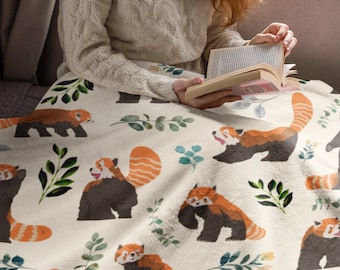 May include: A white throw blanket with a red panda print. The blanket features a repeating pattern of red pandas, leaves, and flowers on a cream background.