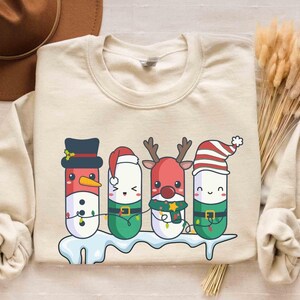 Op de afbeelding: Zandkleurige sweatshirt met een feestelijk ontwerp van vier stripfiguren verkleed als kerstfiguren: een sneeuwman, de kerstman, een rendier en een elf. Elk personage is ontworpen om op een capsule te lijken. Het ontwerp bevat kleurrijke details en een sneeuwbasis.