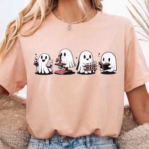 May include: A light pink t-shirt with a graphic of four ghosts dressed in different costumes. The ghosts are holding a potion, a microscope, a musical instrument, and a book. The text "heather peach" is visible in the upper right corner of the image.