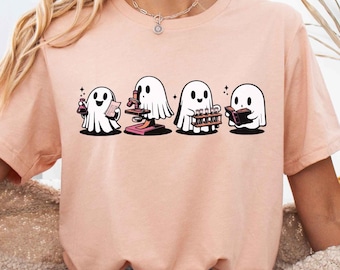 Può includere: Una maglietta rosa chiaro con una grafica di quattro fantasmi vestiti con costumi diversi. I fantasmi tengono una pozione, un microscopio, uno strumento musicale e un libro. Il testo "heather peach" è visibile nell'angolo in alto a destra dell'immagine.