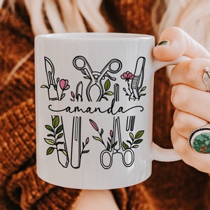 Könnte beinhalten: Weiße Keramiktasse mit einer schwarzen Strichzeichnung von chirurgischen Instrumenten und floralen Akzenten. Der Name "amanda" ist in einer Schreibschrift geschrieben. Die Tasse wird von einer Person gehalten.